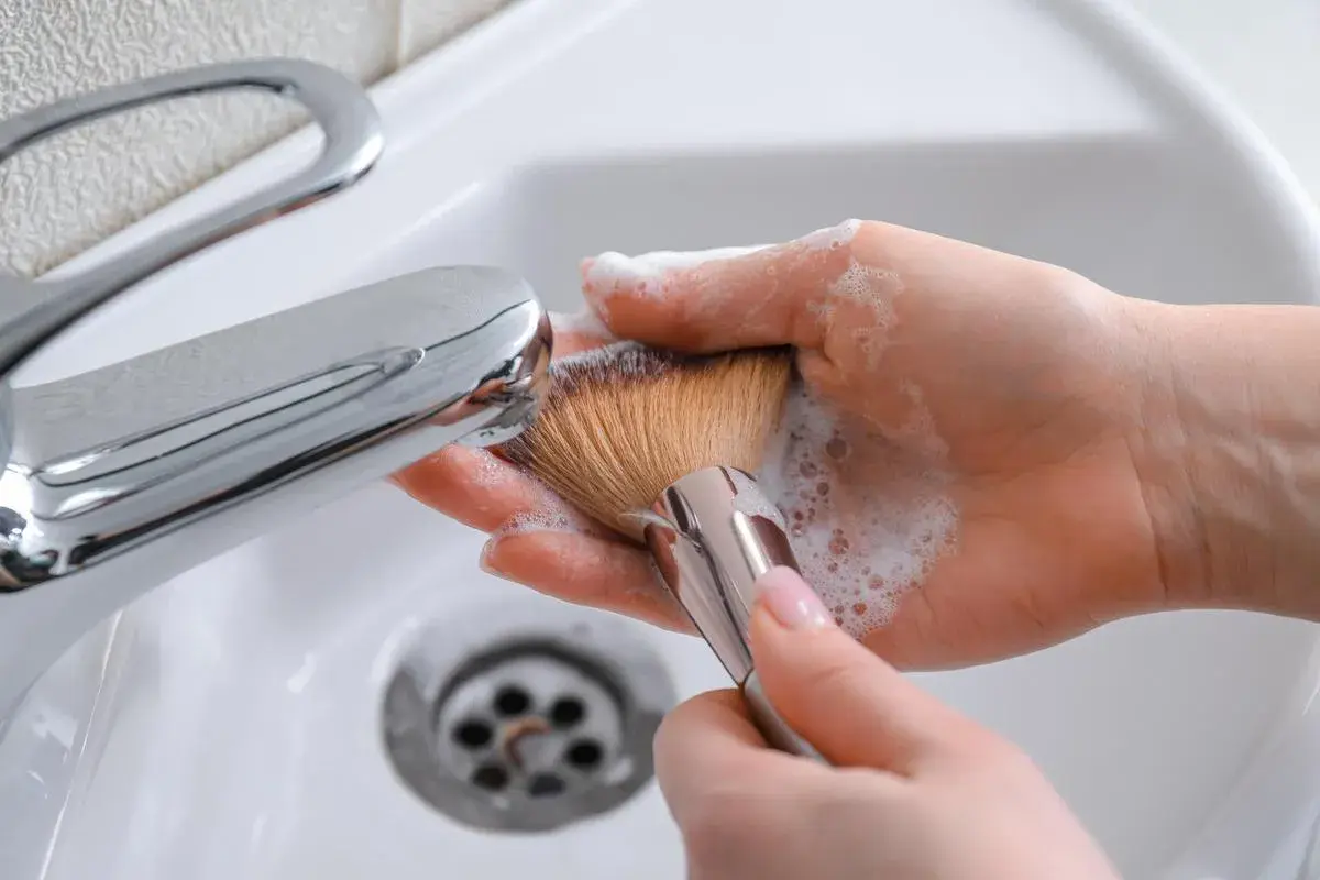 Manos limpiando brochas de maquillaje con espuma y agua bajo un grifo cromado en un lavabo blanco.