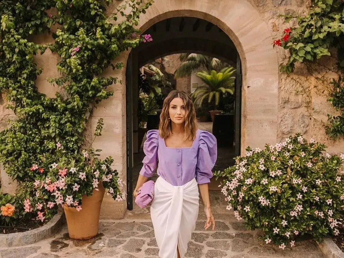 Mujer con ropa que combina color lila claro, un top abullonado y falda blanca, paseando entre vegetación exuberante.