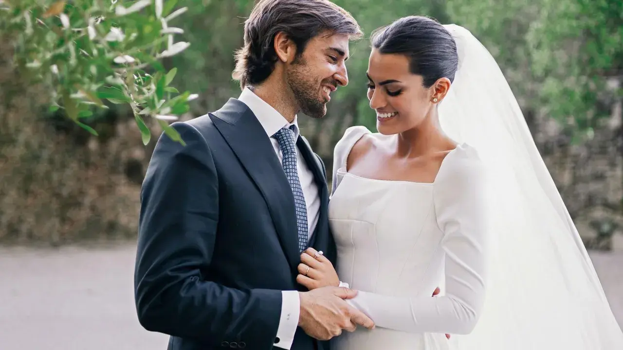 Marta Lozano y su esposo, radiantes en su boda, comparten una mirada cómplice y sonrisas. Él, con traje oscuro y corbata, la abraza tiernamente.