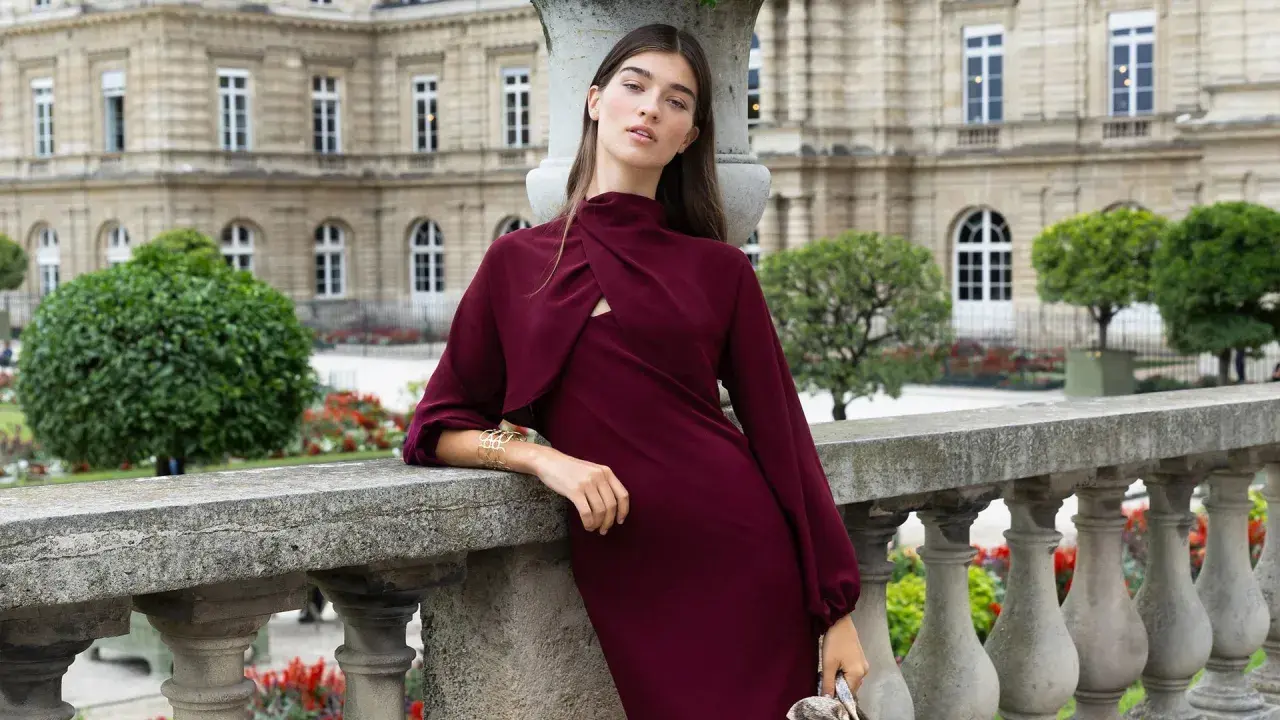 Mujer con un elegante vestido granate, apoyada en una barandilla de piedra, con un edificio histórico y jardines de fondo.