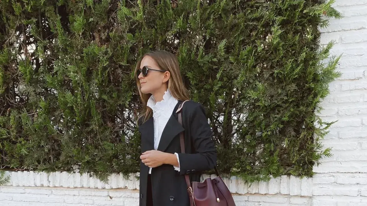 Mujer con elegante outfit para Semana Santa, abrigo negro, blusa blanca con volantes y bolso burdeos.