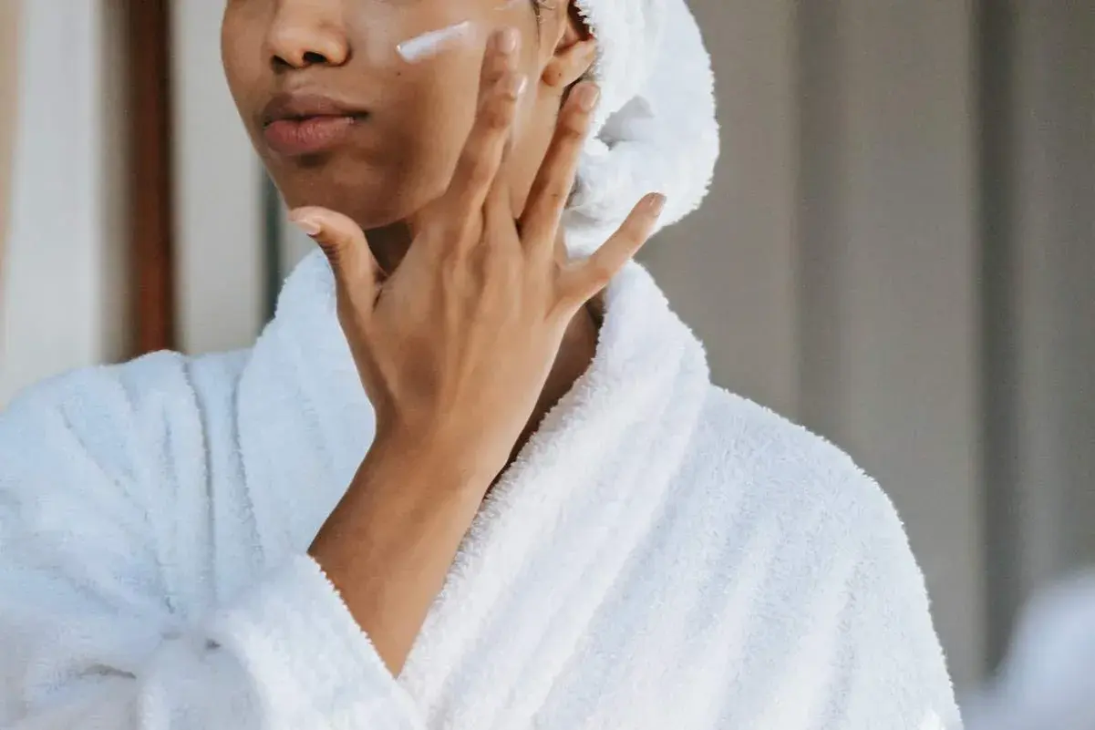 Mujer con bata blanca y toalla en la cabeza aplicándose crema en la mejilla, un gesto de cuidado para la piel con colágeno.