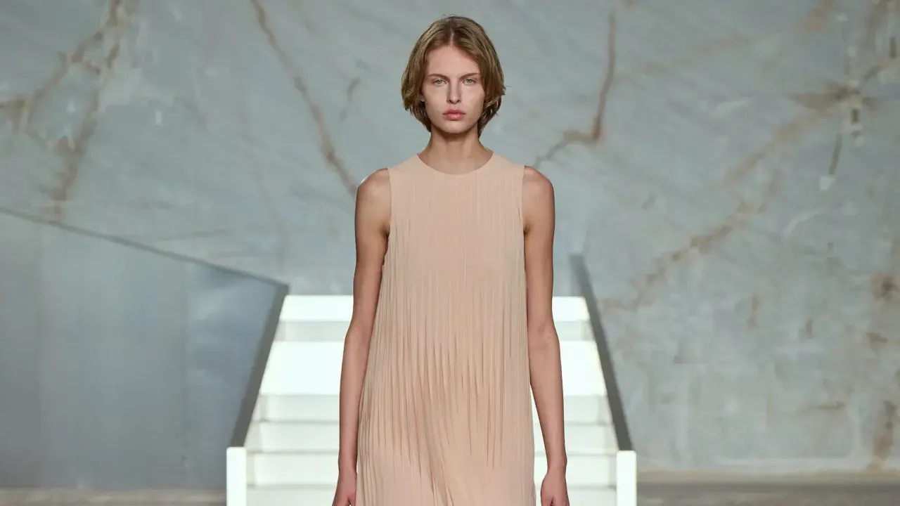 Modelo con vestido plisado color melocotón, en un escenario minimalista con escaleras blancas y fondo de mármol.