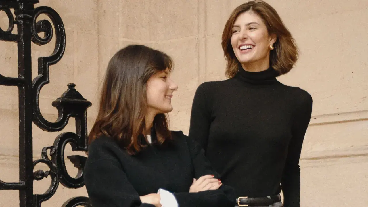 María de la Orden y su amiga ríen juntas frente a una reja ornamental.