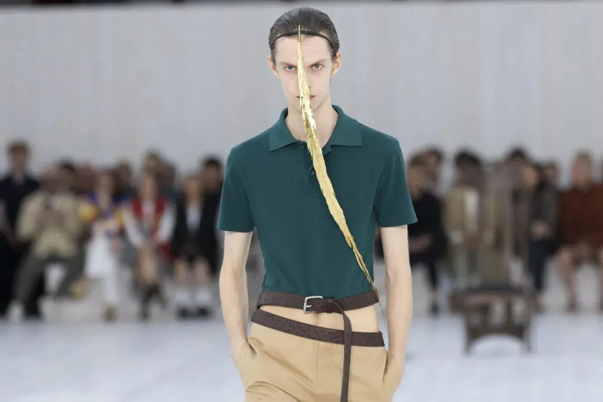 Enzo Vogrincic, modelo con un polo verde, pantalones caqui y un adorno dorado en la cara, desfila ante un público difuminado.