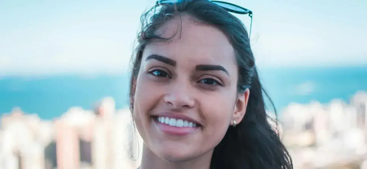 Sonrisa radiante de una joven con rostro ovalado, cabello oscuro y gafas de sol en la cabeza. Fondo de ciudad y mar.