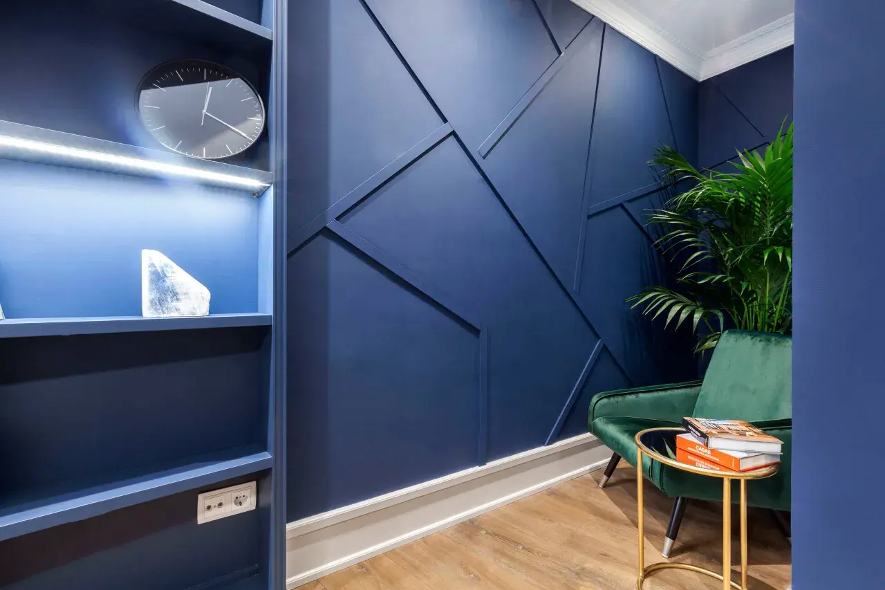 Un sillón verde menta contrasta con la pared azul oscuro. Una planta y libros descansan sobre una mesa dorada.