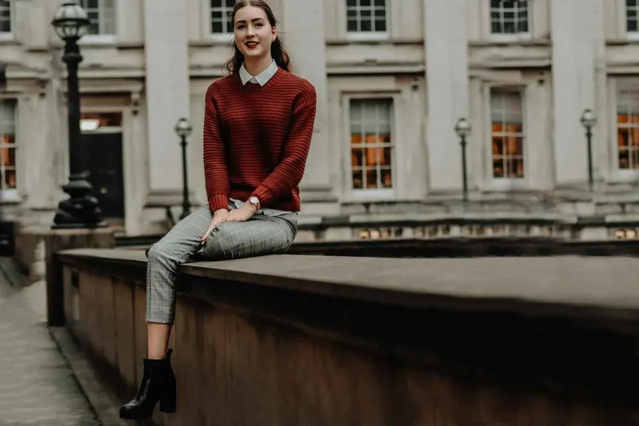 Chica con estilos de ropa modernos: jersey de punto rojo, camisa blanca, pantalones de cuadros y botines negros, sentada frente a un edificio clásico.