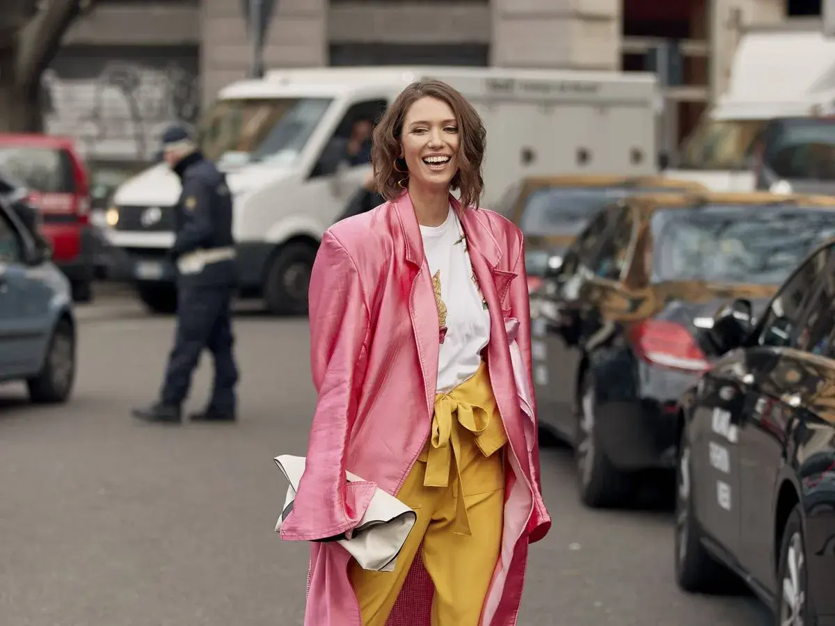 Mujer sonriente con abrigo rosa y pantalones amarillos, demostrando los **colores que combinan con amarillo** en un día urbano.