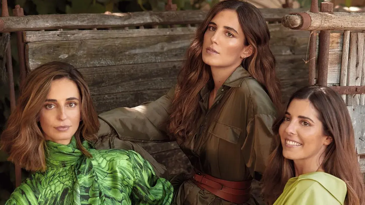 Belen Corsini y dos mujeres más, vestidas con ropa de moda, posan sentadas en un banco de madera rústico.