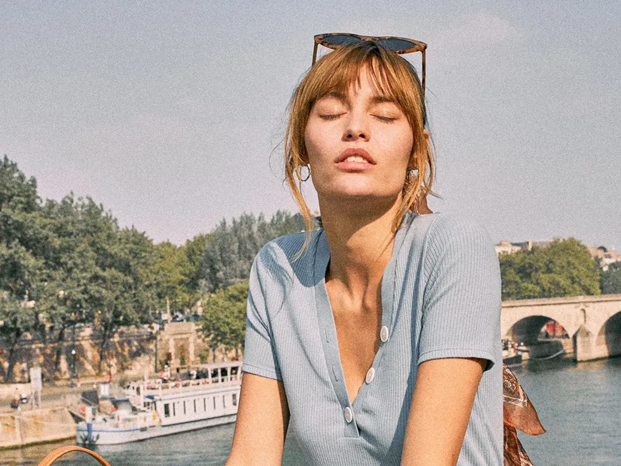 Mujer con gafas de sol en la cabeza y un pañuelo con estampado vichy, disfrutando del sol junto a un río con un barco y un puente al fondo.
