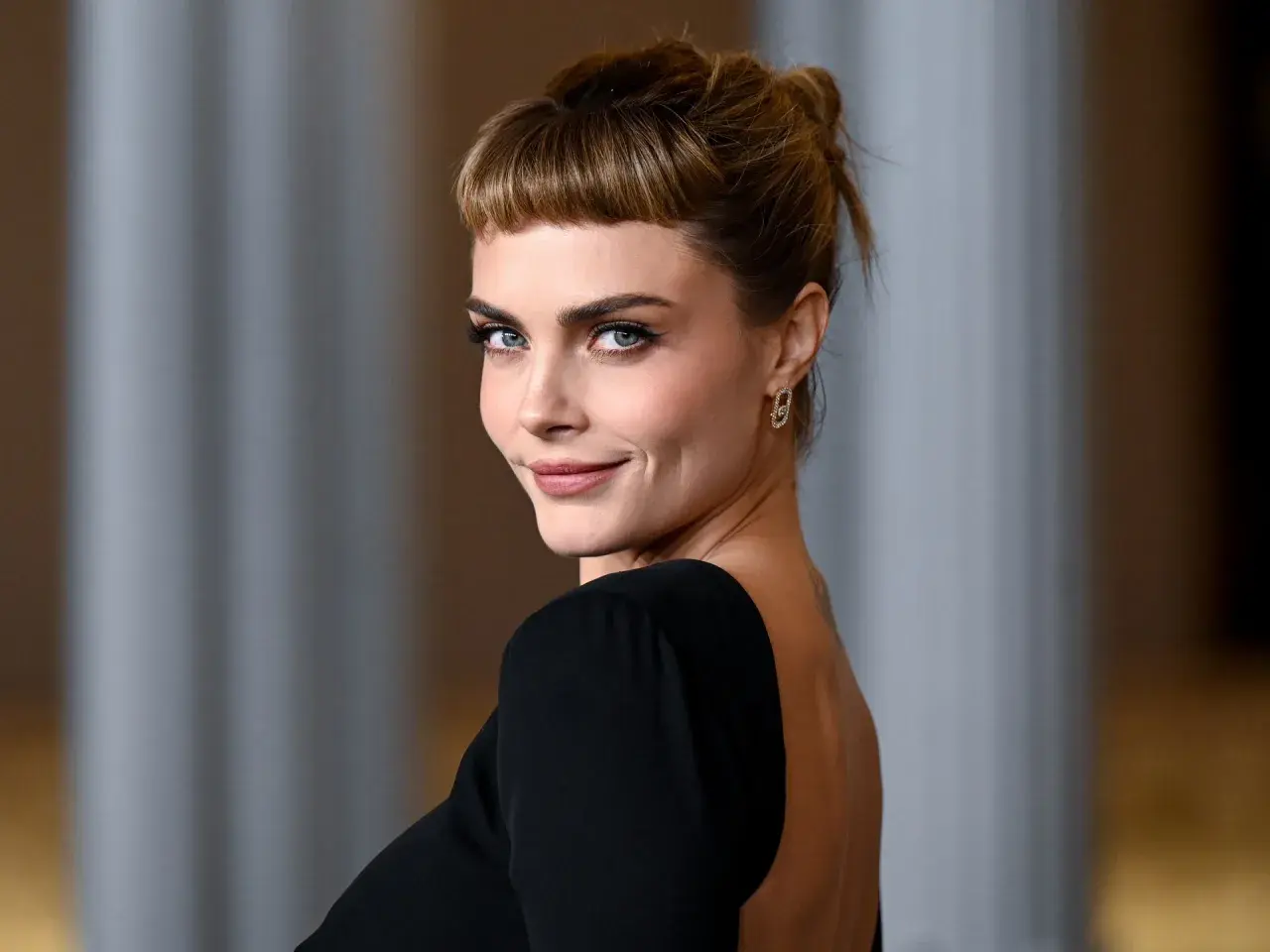 Cara Delevingne, con su mirada penetrante y un peinado recogido con flequillo, luce un elegante vestido negro de espalda descubierta.