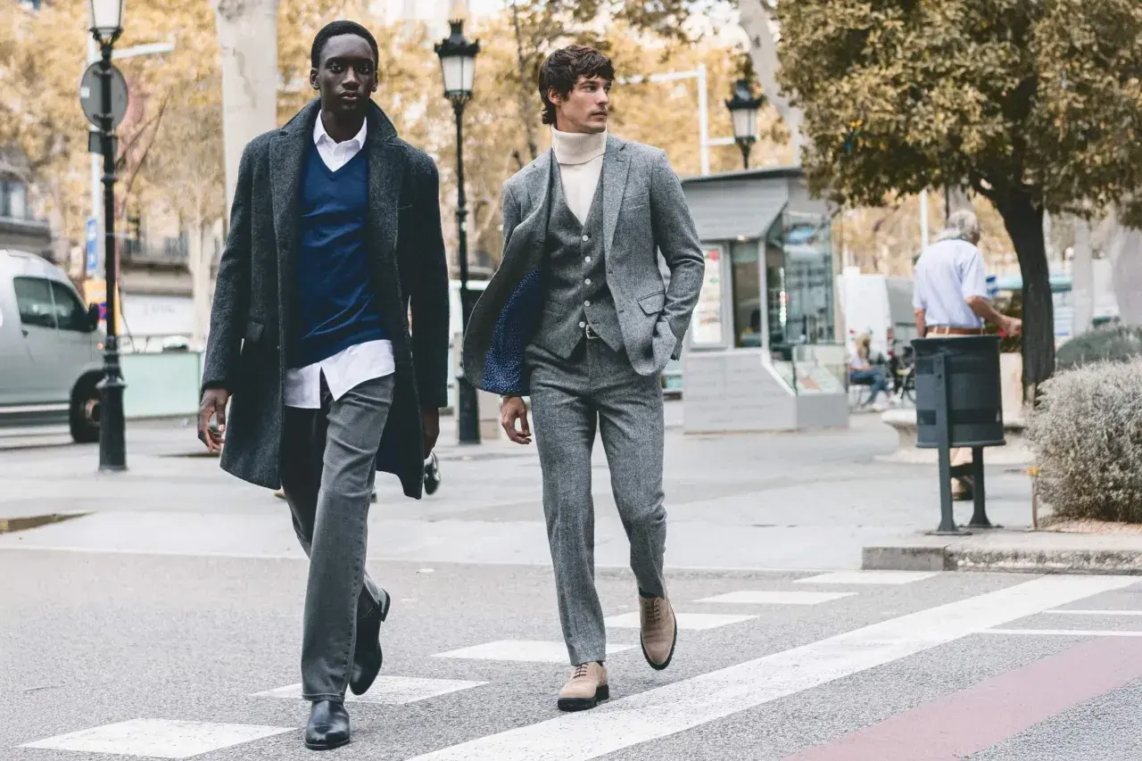 Dos hombres cruzan una calle, uno con abrigo y jersey azul, el otro con traje gris. Representan la moda slow, atemporal y consciente.