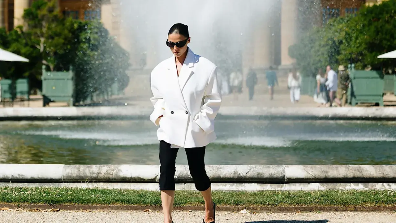 Mujer con un elegante outfit con leggins negro y blazer blanco, paseando cerca de una fuente.