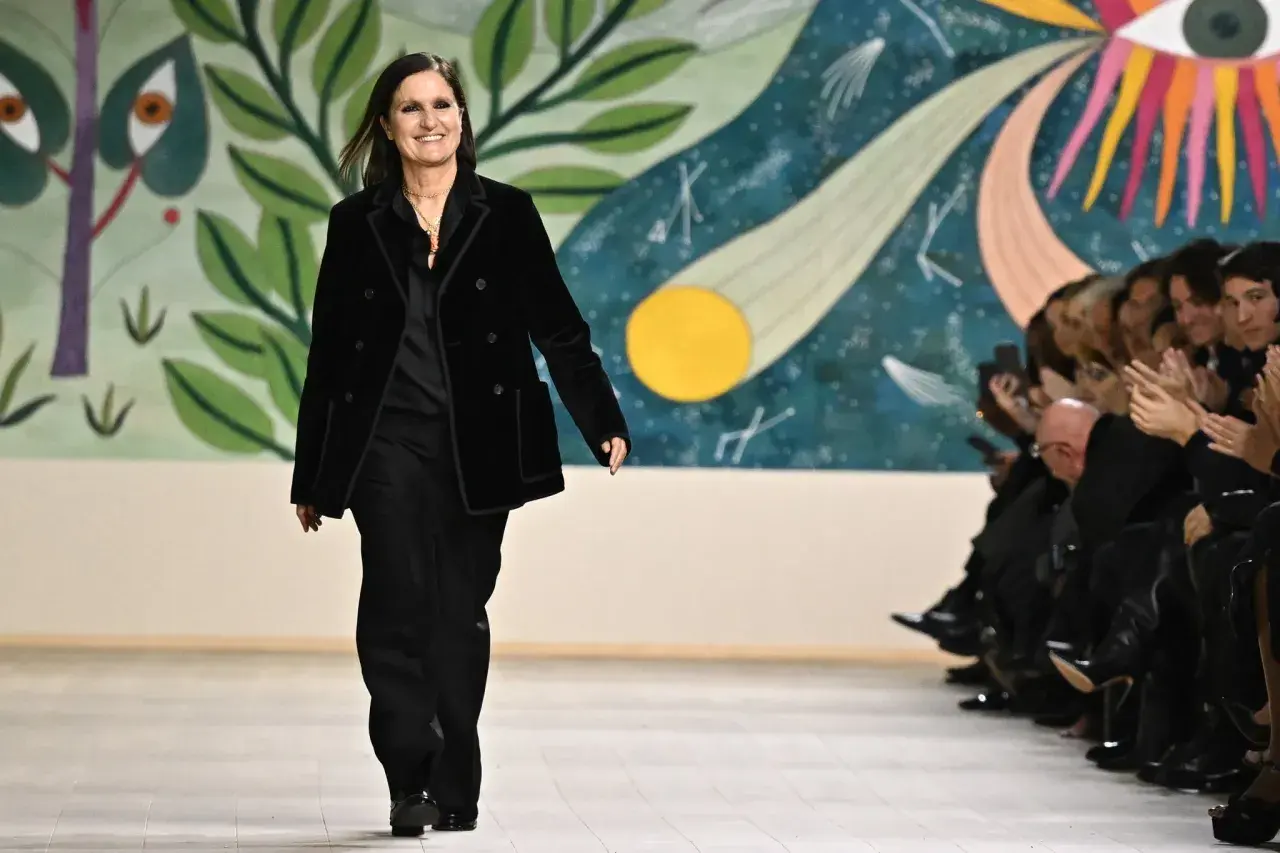 Maria Grazia Chiuri camina en la pasarela con un traje negro, sonriendo ante el público aplaudiendo.