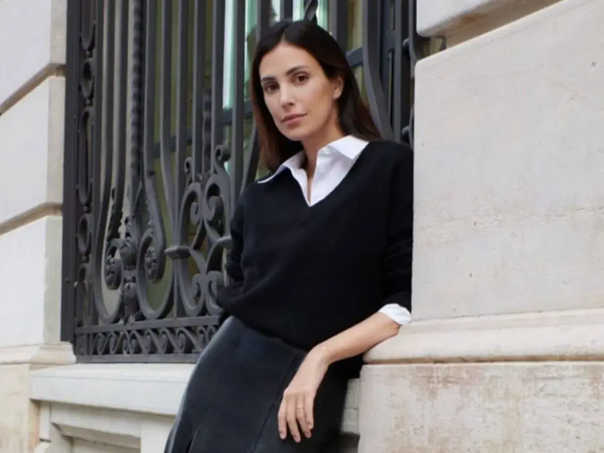 Sassa de Osma, elegante con jersey negro y falda de cuero, apoyada en una columna de piedra junto a una reja ornamental.
