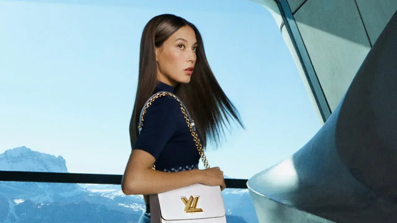 Eileen Gu, con su bolso Louis Vuitton, posa con elegancia frente a un paisaje montañoso y un cielo azul.