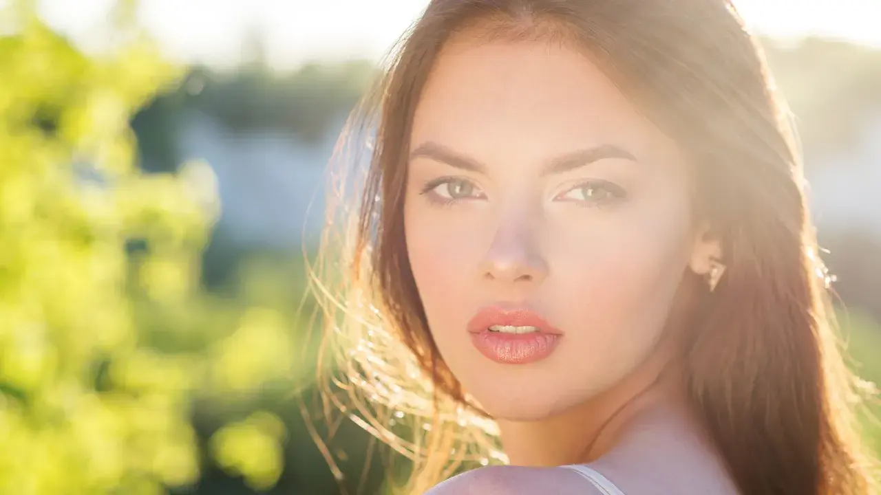 Una joven con un brillo natural, un verdadero glow up, mira a la cámara con el sol iluminando su rostro y el fondo verde desenfocado.