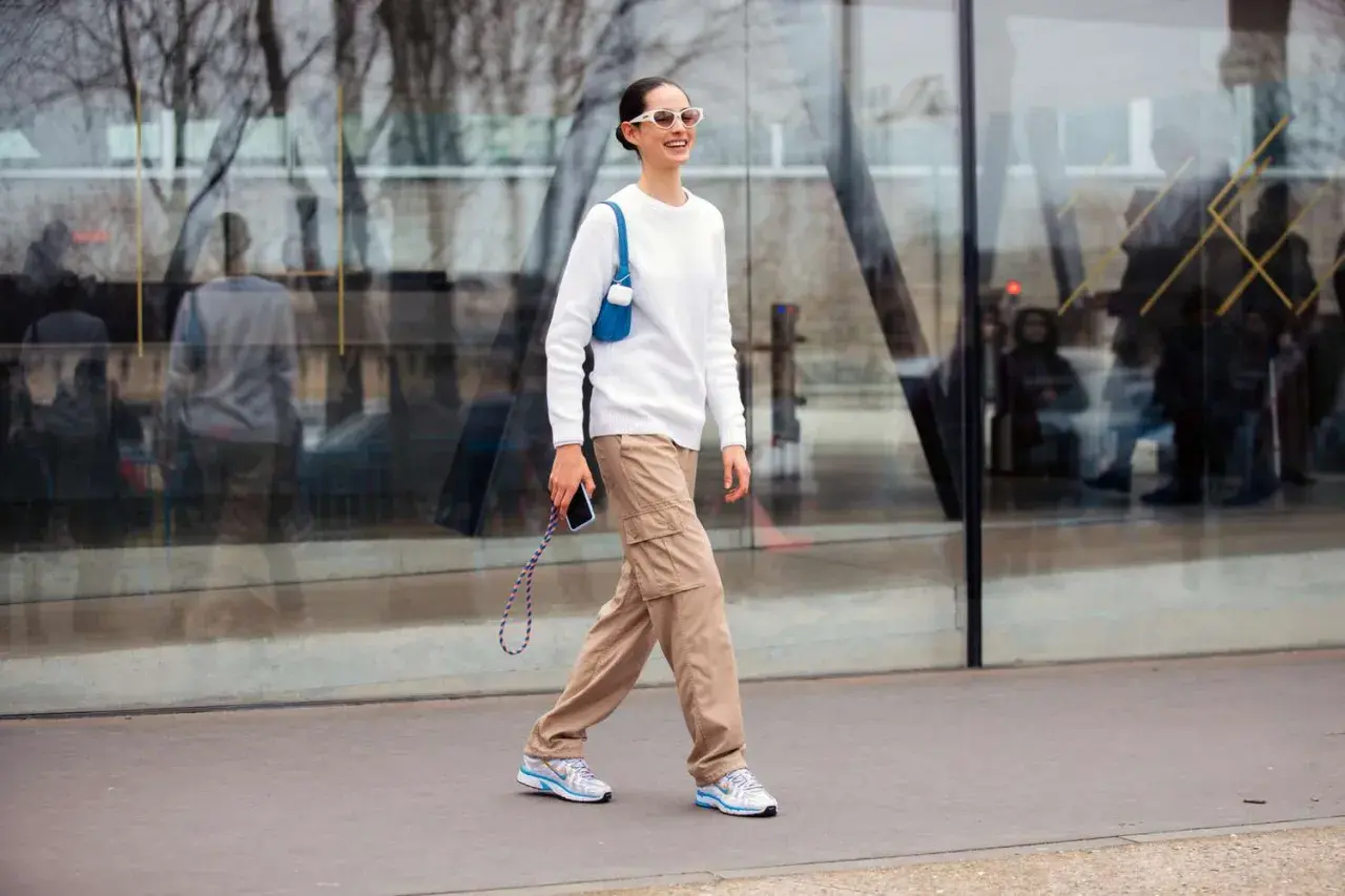 Mujer sonriente camina con un suéter blanco y un pantalón beige de carga, luciendo gafas de sol blancas y un bolso azul.