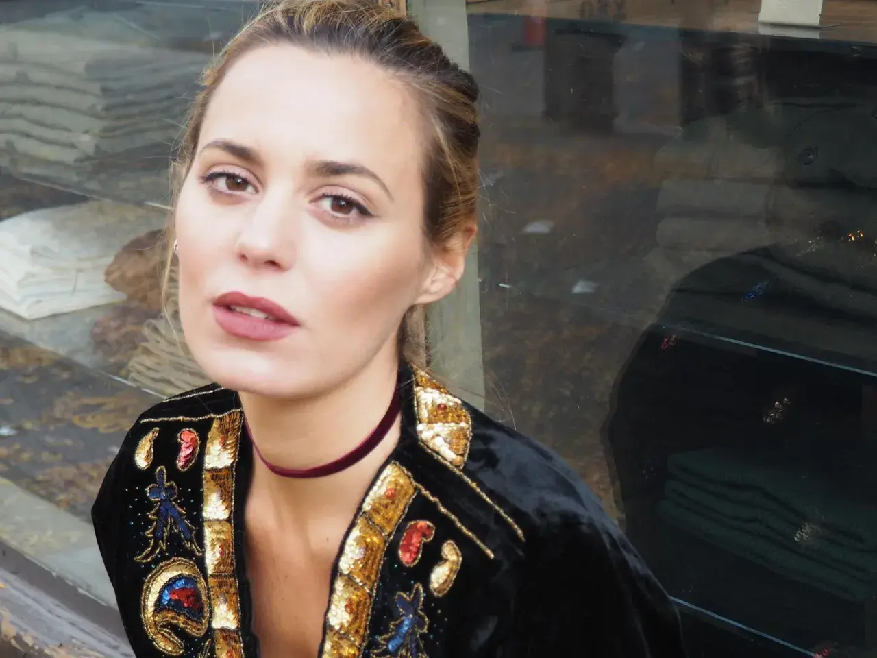 Claudia Ortiz Domecq, con un choker de terciopelo y chaqueta bordada, posa con mirada intensa frente a un escaparate.