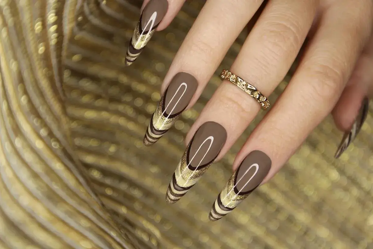 Uñas marrones tendencia con diseño de líneas doradas y brillo. Un anillo dorado adorna el dedo.