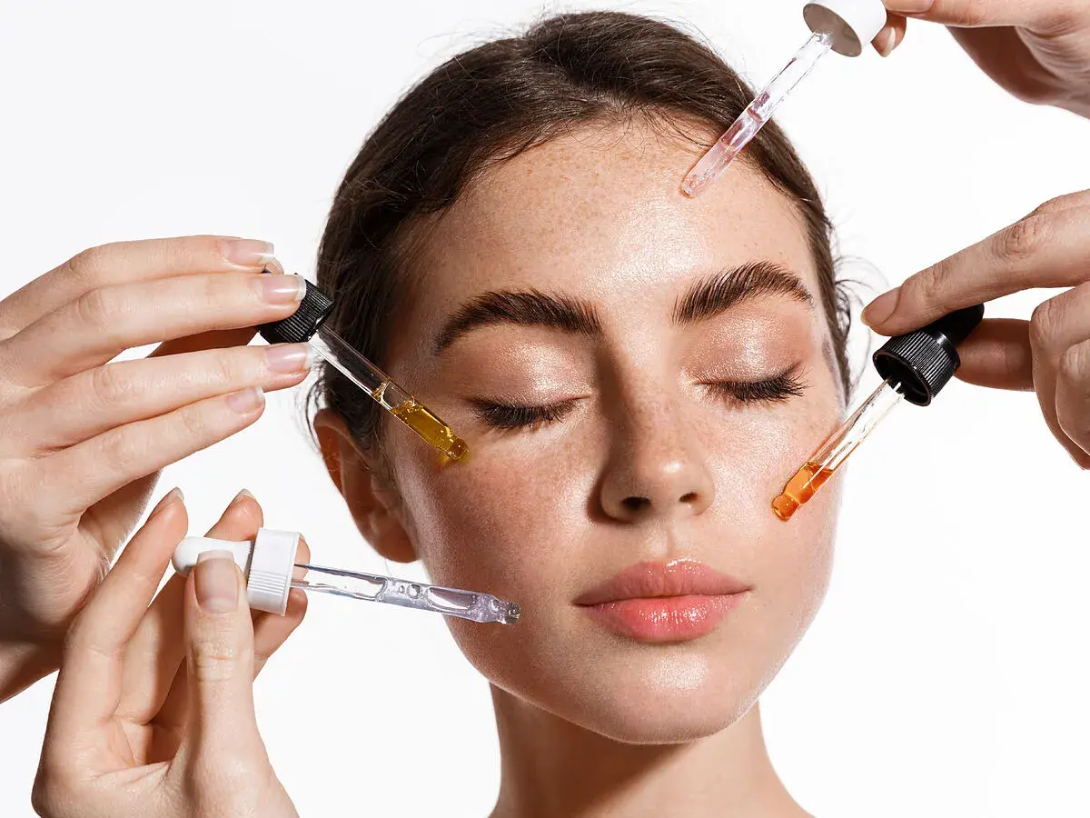 Manos aplican sueros con goteros en el rostro de una mujer. El brillo en su piel sugiere el poder de la vitamina C para la cara.