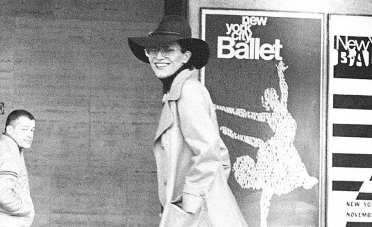 Elsa Peretti, con sombrero y gafas, sonríe frente a un cartel del New York City Ballet.