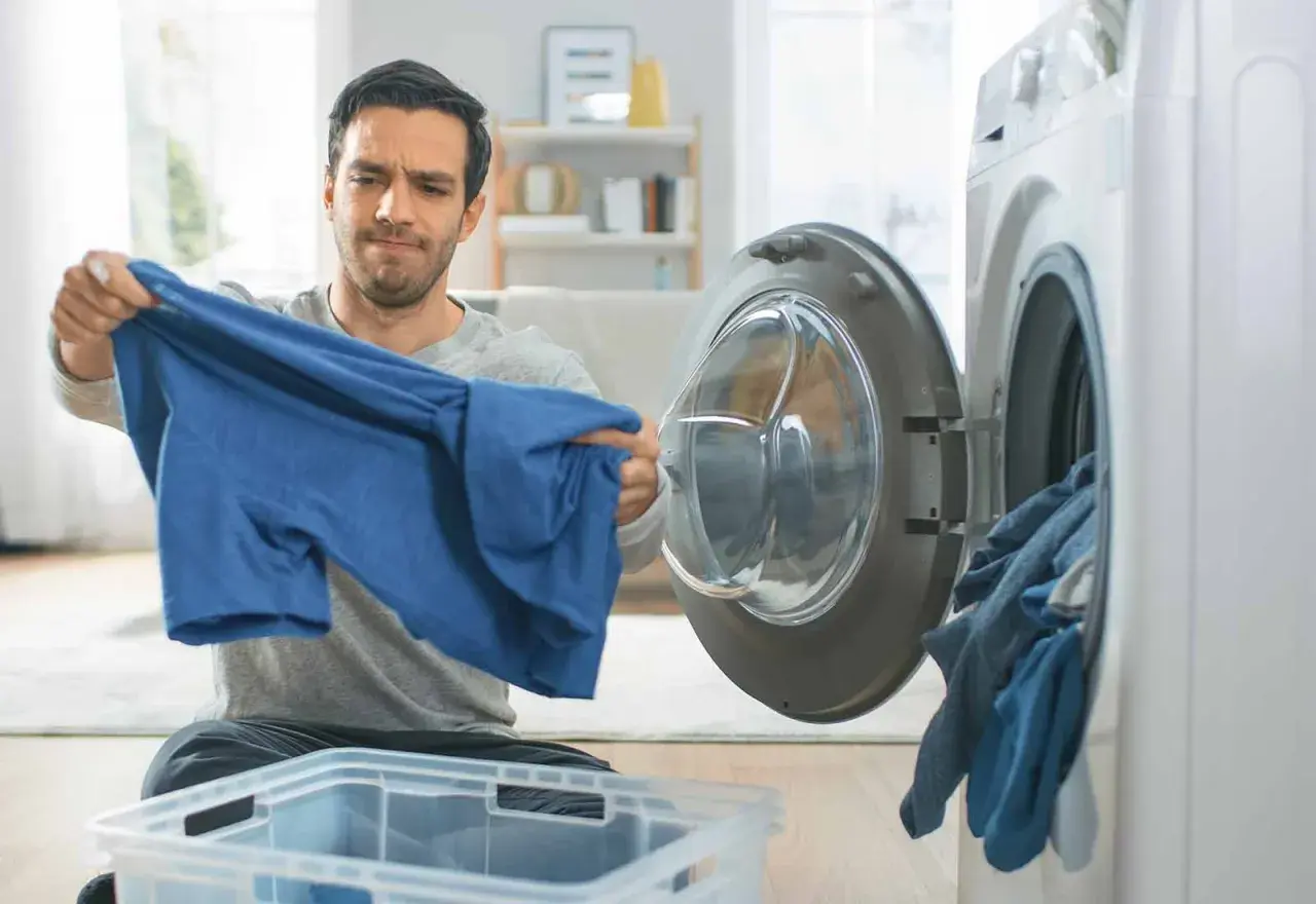Hombre mira con preocupación una camiseta azul, parece que la viscosa encoge. La lavadora está abierta con ropa dentro.