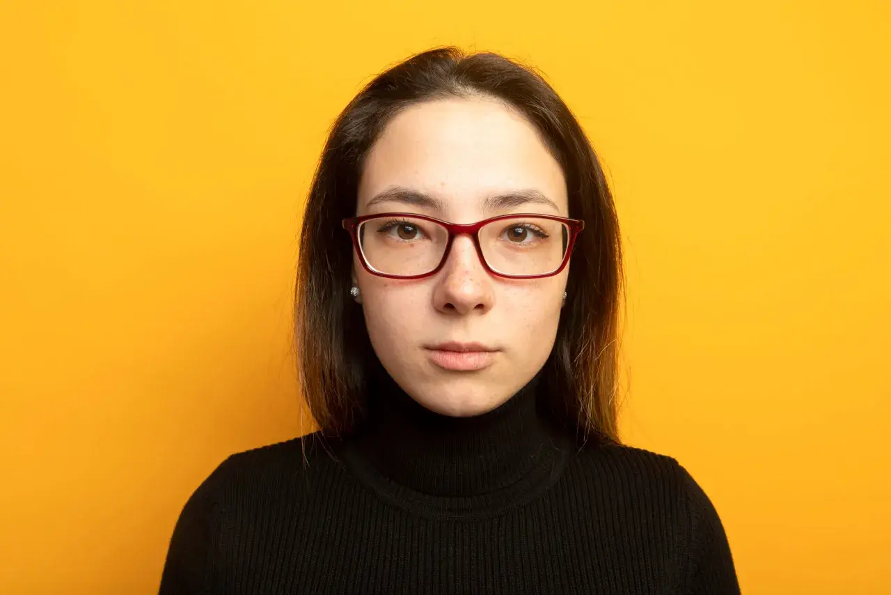 Joven con gafas de cara alargada y estrecha, pecas y jersey negro, sobre fondo amarillo.