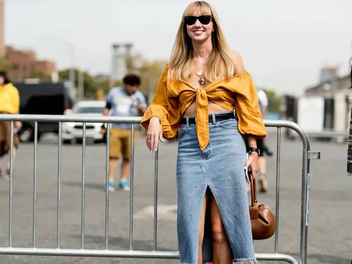 Mujer sonriente con un outfit falda vaquera larga, top amarillo anudado y bolso marrón.