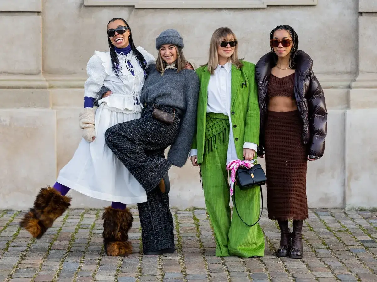 Cuatro amigas, con estilos audaces, posan juntas. Una lleva botas peludas, otra un traje verde vibrante, y la tercera un conjunto marrón. ¡Estilo como el de Odessa A'zion!