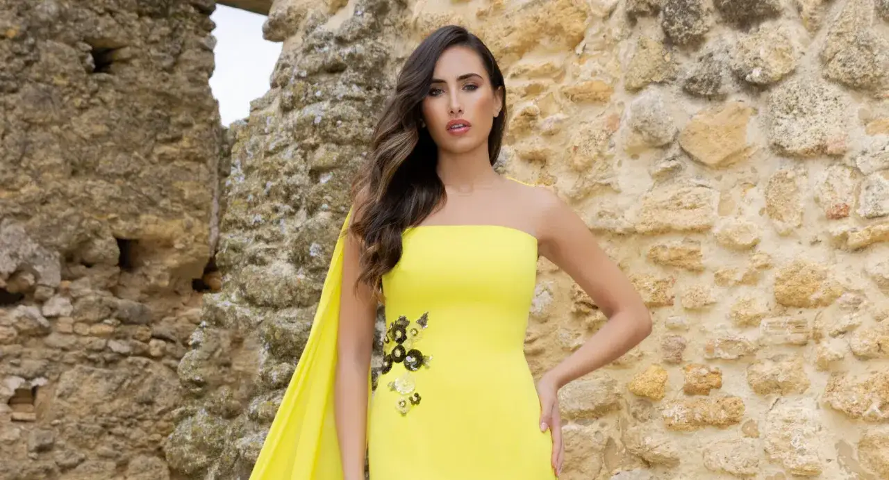 Mujer con vestido amarillo vibrante, escote palabra de honor con detalles florales, posando frente a una pared de piedra antigua.