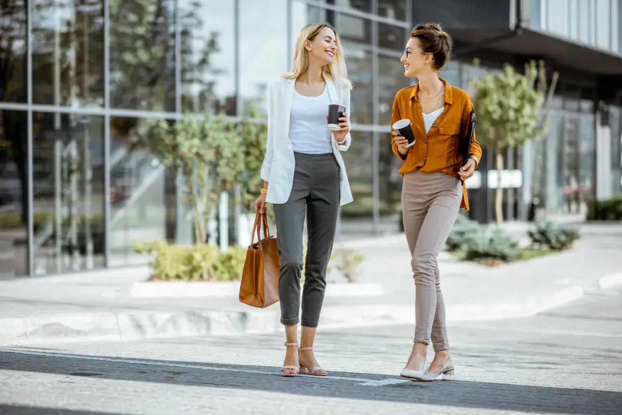 Dos mujeres con atuendo business casual caminan y conversan frente a un edificio moderno, cada una con un café en mano.