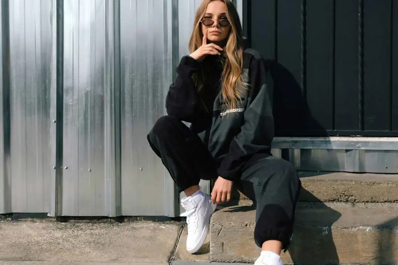 Chica con gafas de sol y ropa deportiva, sentada en escalones de hormigón. Su estilo es un ejemplo de la estética urbana que es.