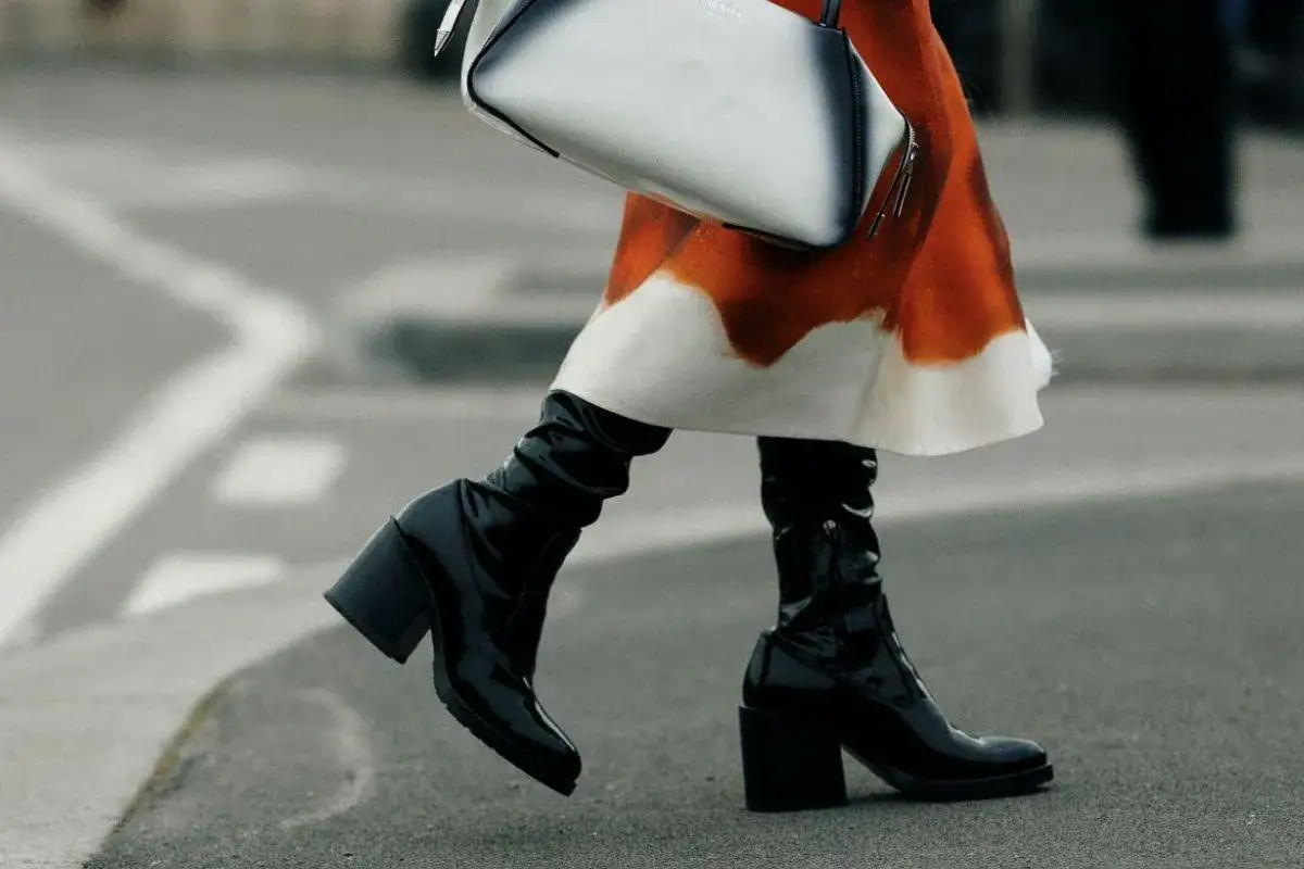 Mujer con falda naranja y blanca y unas botas negras brillantes con tacón grueso, caminando por la calle.