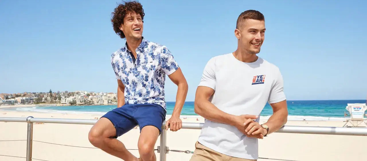Dos hombres en la playa. Uno lleva una camisa estampada y un outfit de pantalon corto hombre azul. El otro, camiseta blanca y bermudas.