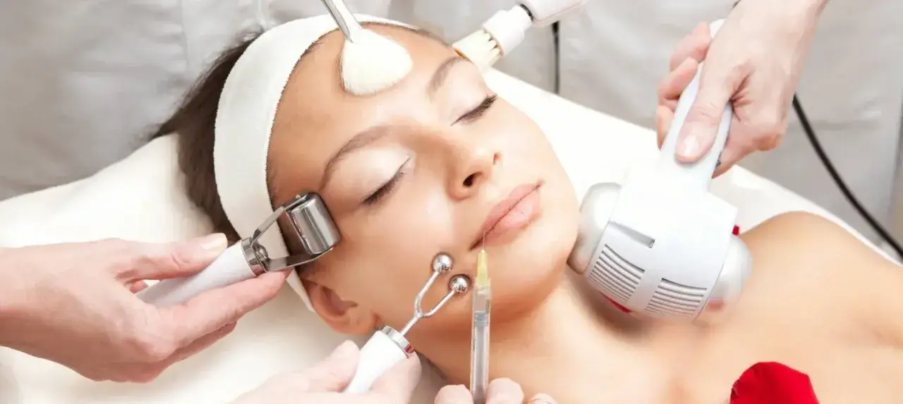 Mujer recibiendo un tratamiento facial con varios dispositivos, incluyendo un rodillo, una jeringa y un cepillo.