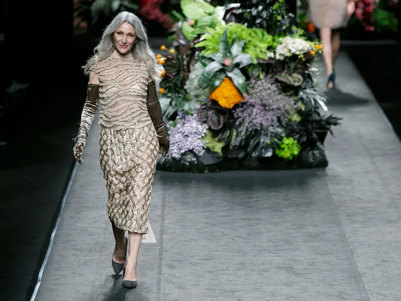 Una modelo con cabello plateado desfila con un vestido de lentejuelas y guantes largos, luciendo como un pino montesdeoca en un jardín exuberante.