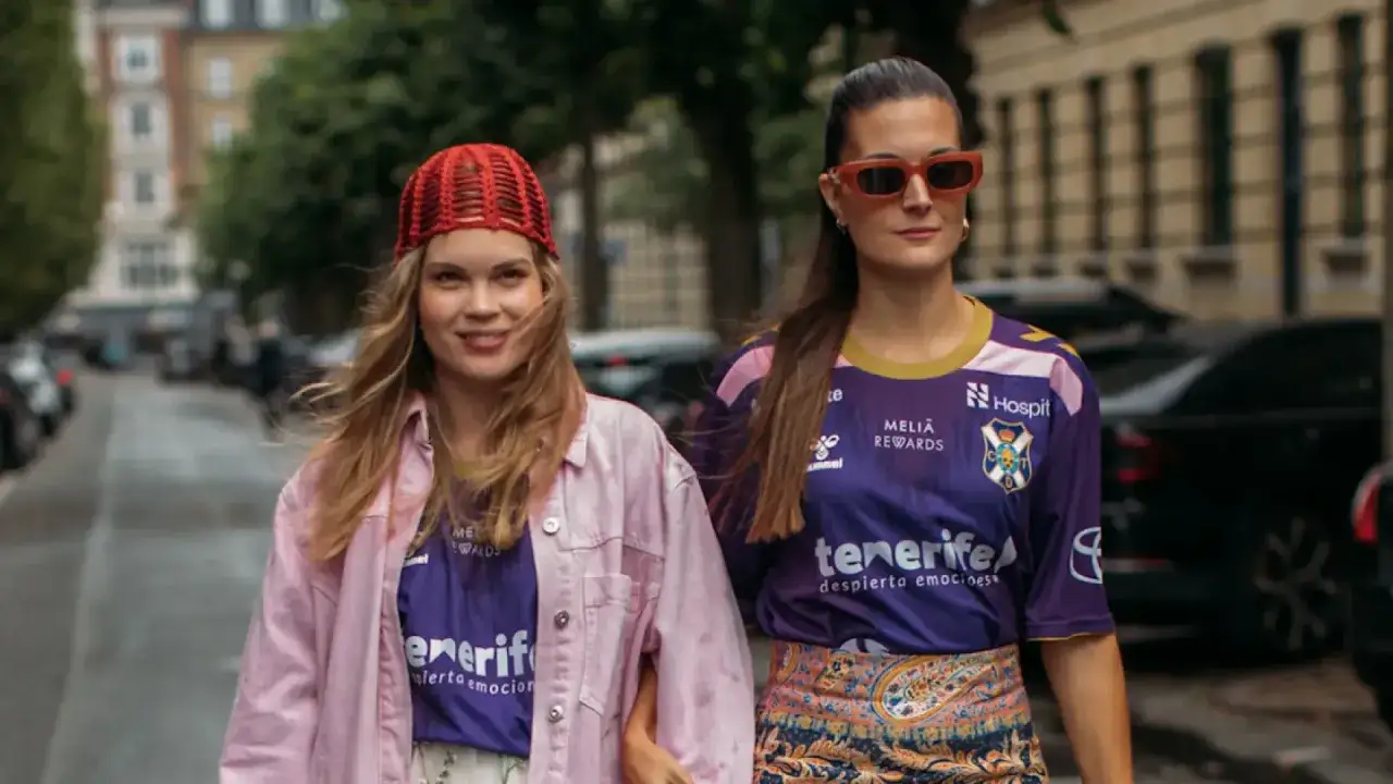 Dos amigas pasean con estilo blokecore, una con gorro rojo y chaqueta rosa, la otra con gafas naranjas y camiseta de fútbol.