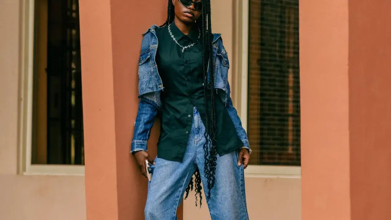 Chica con trenzas largas y gafas de sol, vestida con camisa negra, chaqueta vaquera y vaqueros anchos. Su estilo es muy aesthetic.