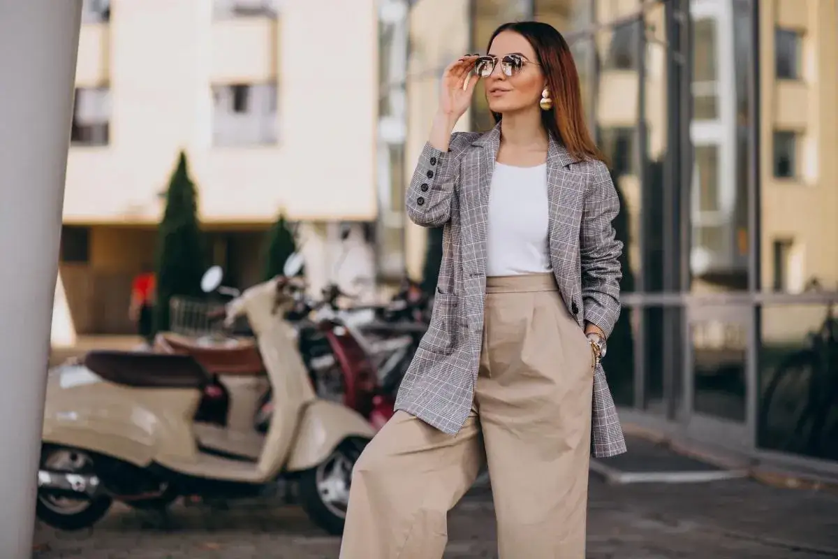 Mujer con traje de cuadros y pantalones anchos, gafas de sol y pendientes dorados, encarnando el lujo silencioso junto a una moto clásica.