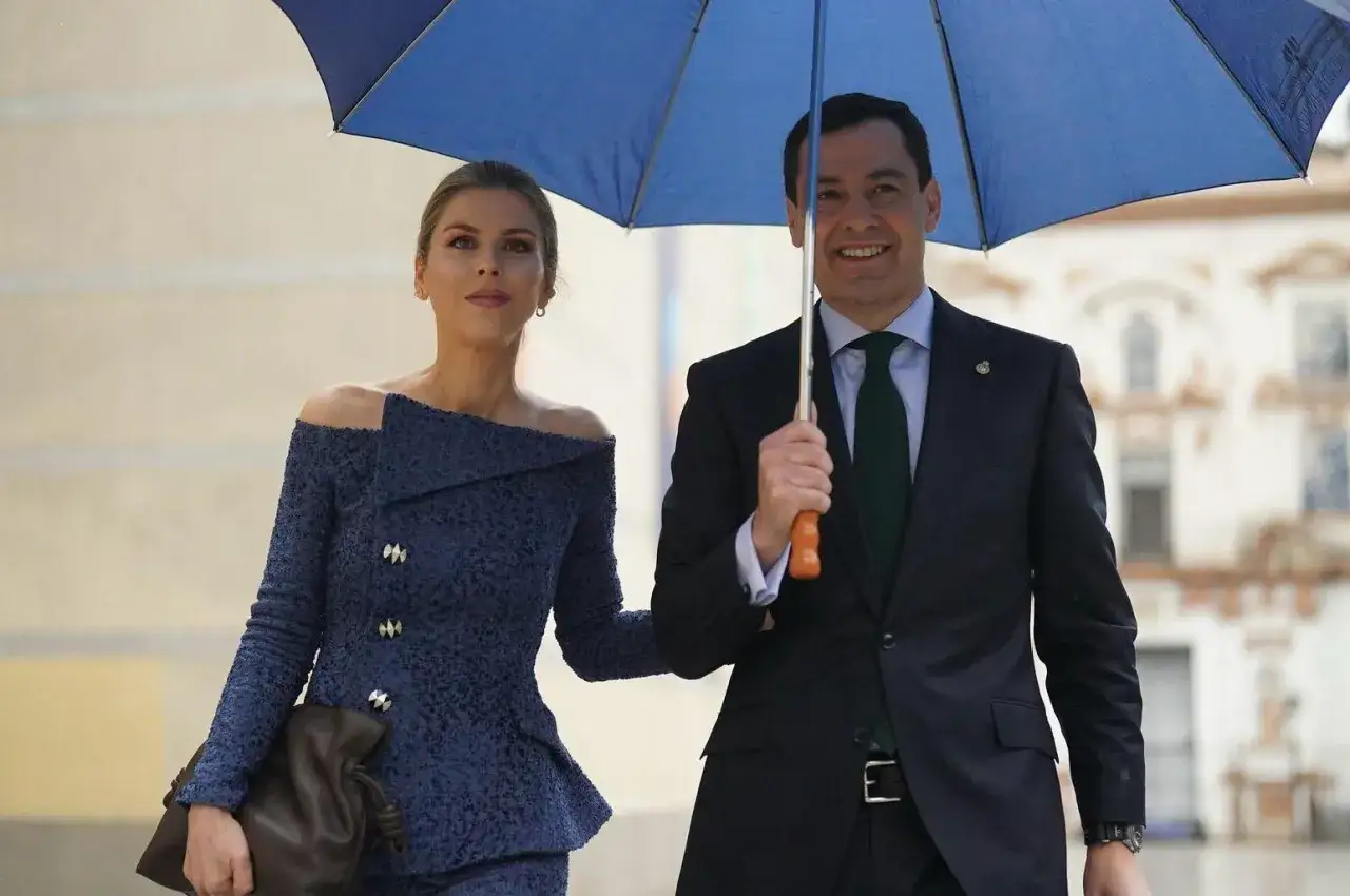 Manuela Villena y un hombre sonríen bajo un paraguas azul. Ella lleva un elegante traje azul, él un traje oscuro.