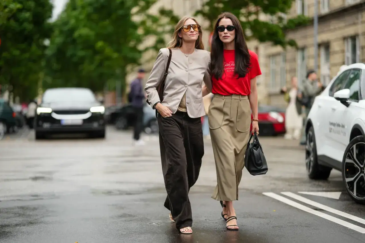 Dos mujeres con looks casuales caminan por la calle. Una lleva un blazer beige y pantalones anchos marrones, la otra un top rojo y falda larga caqui.