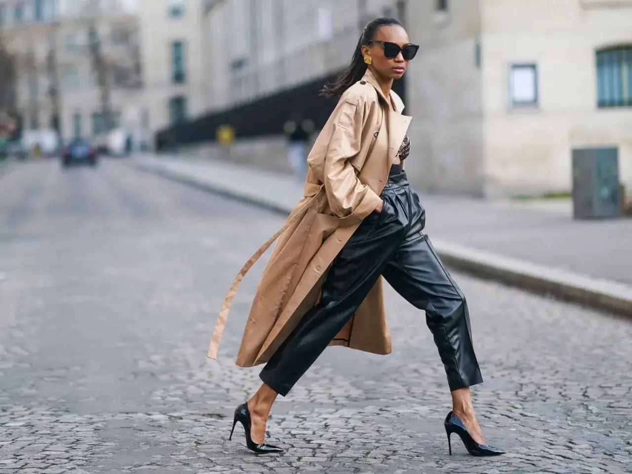 Mujer con estilo parisino, luciendo un trench color camel, pantalones de cuero negros y tacones altos, camina por una calle empedrada.