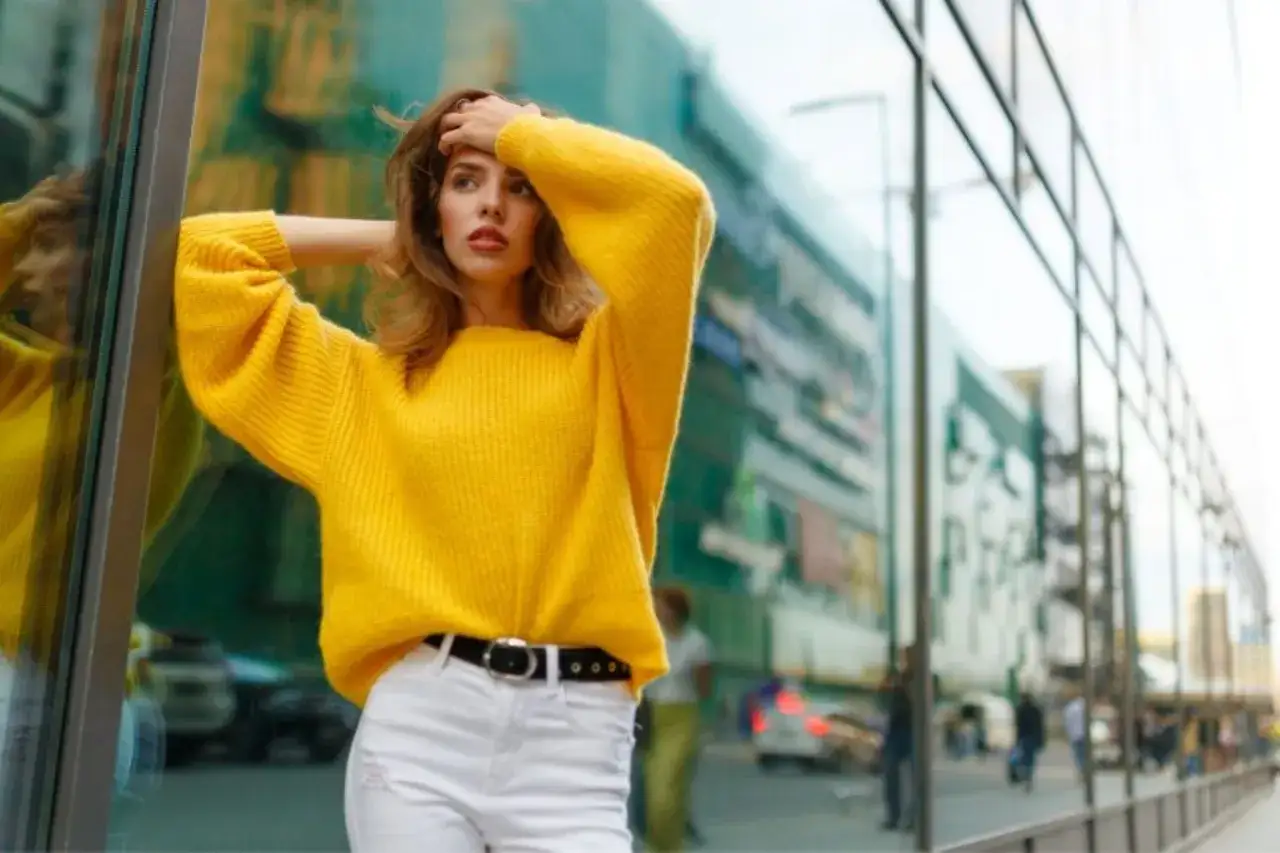 Mujer con suéter amarillo vibrante y pantalones blancos, posando frente a un edificio con reflejos. Los colores amarillo y blanco resaltan.