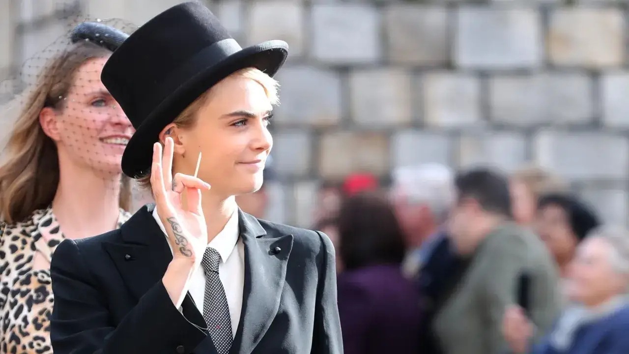 Cara Delevingne, con su estilo tomboy, luce un traje negro y sombrero de copa, saludando con la mano.