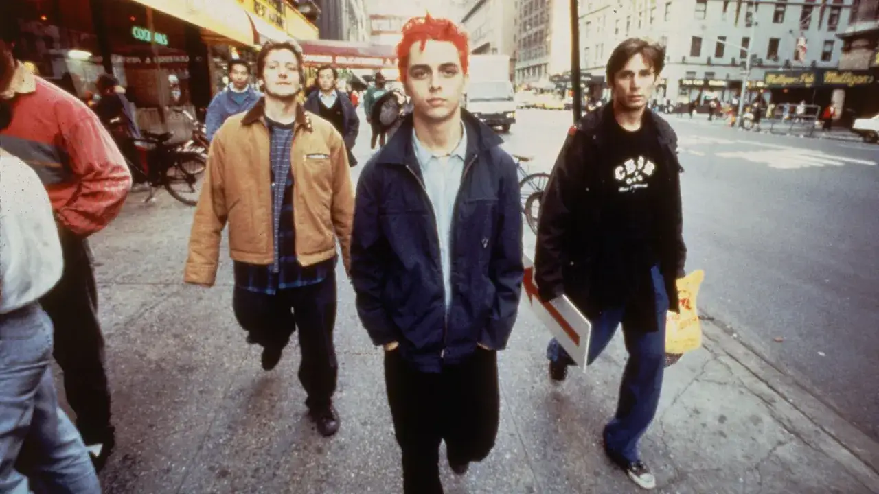 Jóvenes con estilo de los años 90 moda, caminan por la calle. Uno con pelo rojo vibrante, otro con chaqueta marrón y camisa a cuadros, y el tercero con camiseta negra.