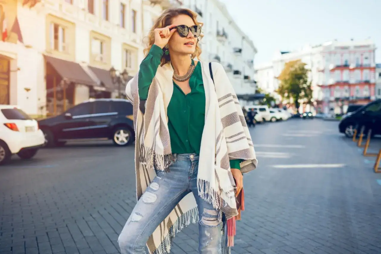 Mujer de 40 años con look casual, gafas de sol, blusa verde, jeans rotos y un poncho a rayas.