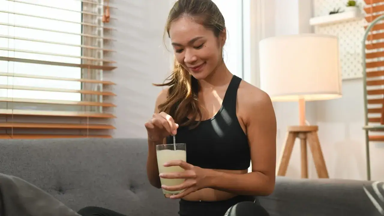 Mujer sonriente prepara bebida de Oslo Skin Lab en un vaso con pajita, sentada en un sofá.