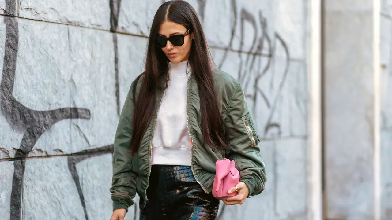 Mujer con gafas de sol y una bomber verde, sostiene un bolso rosa.
